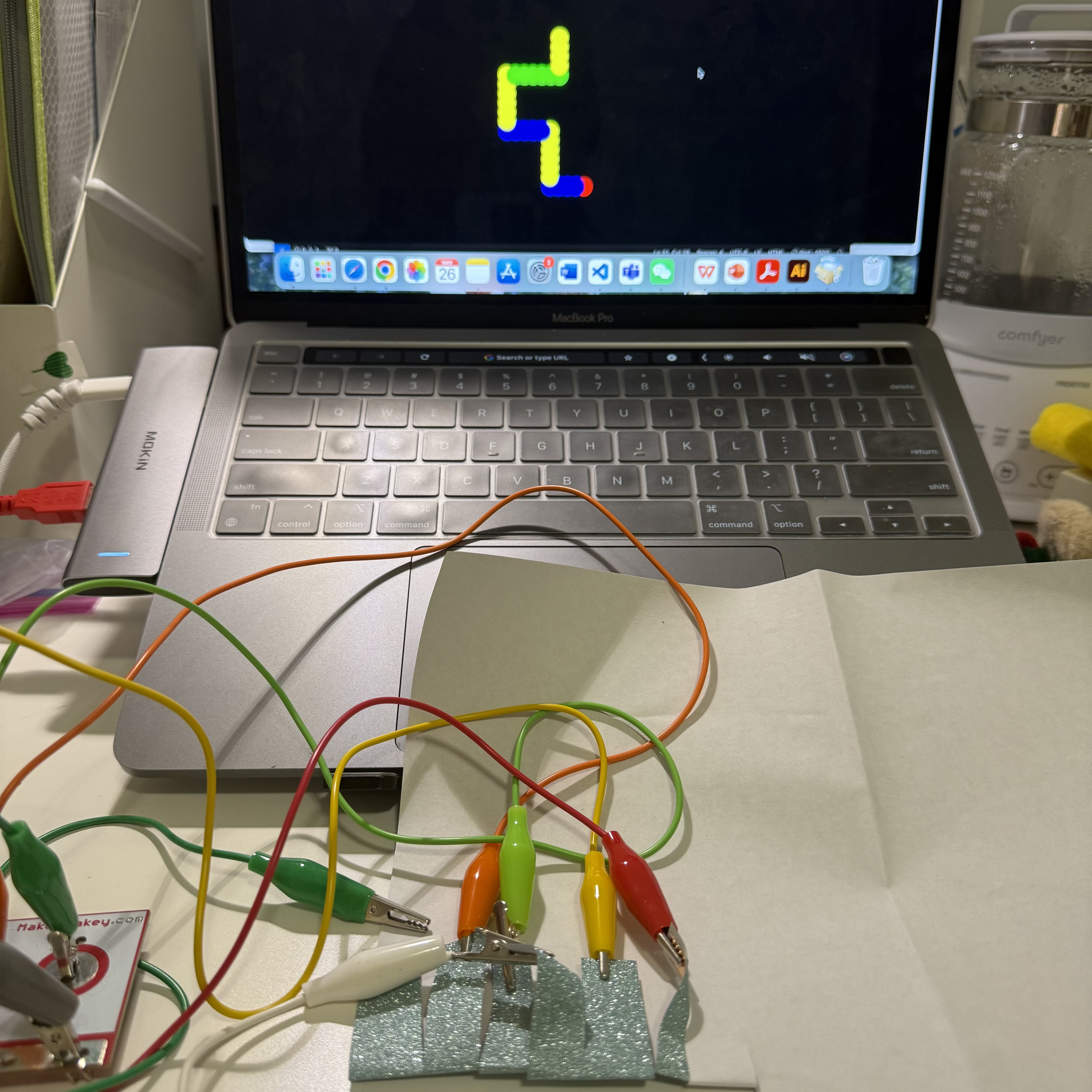 In the setup, there is a Makey Makey board connected to strips of conductive material through colorful alligator clips. The setup is used for creating an interactive electronic project, allowing the connection of everyday objects to computer programs.